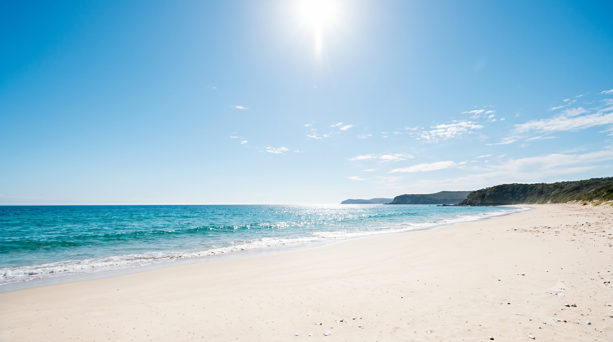 Sun high in a clear blue sky over a quiet Australian beach with calm turquoise ocean and white sand