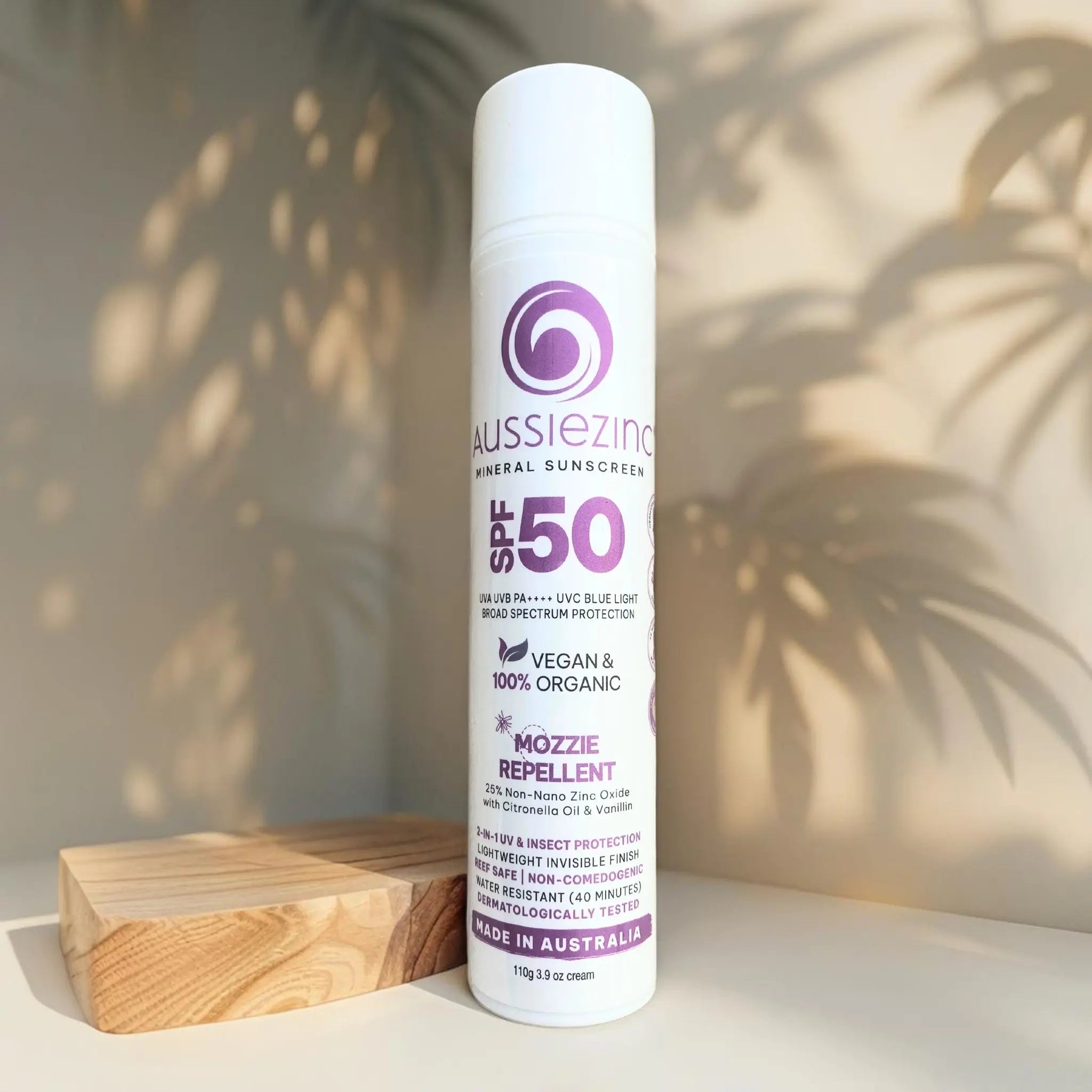 A bottle of AussieZinc 2-in-1 Mozzie Repellent SPF50+ Organic Mineral Zinc Sunscreen placed on a wooden surface with a shadow of a plant leaf on the bottle.