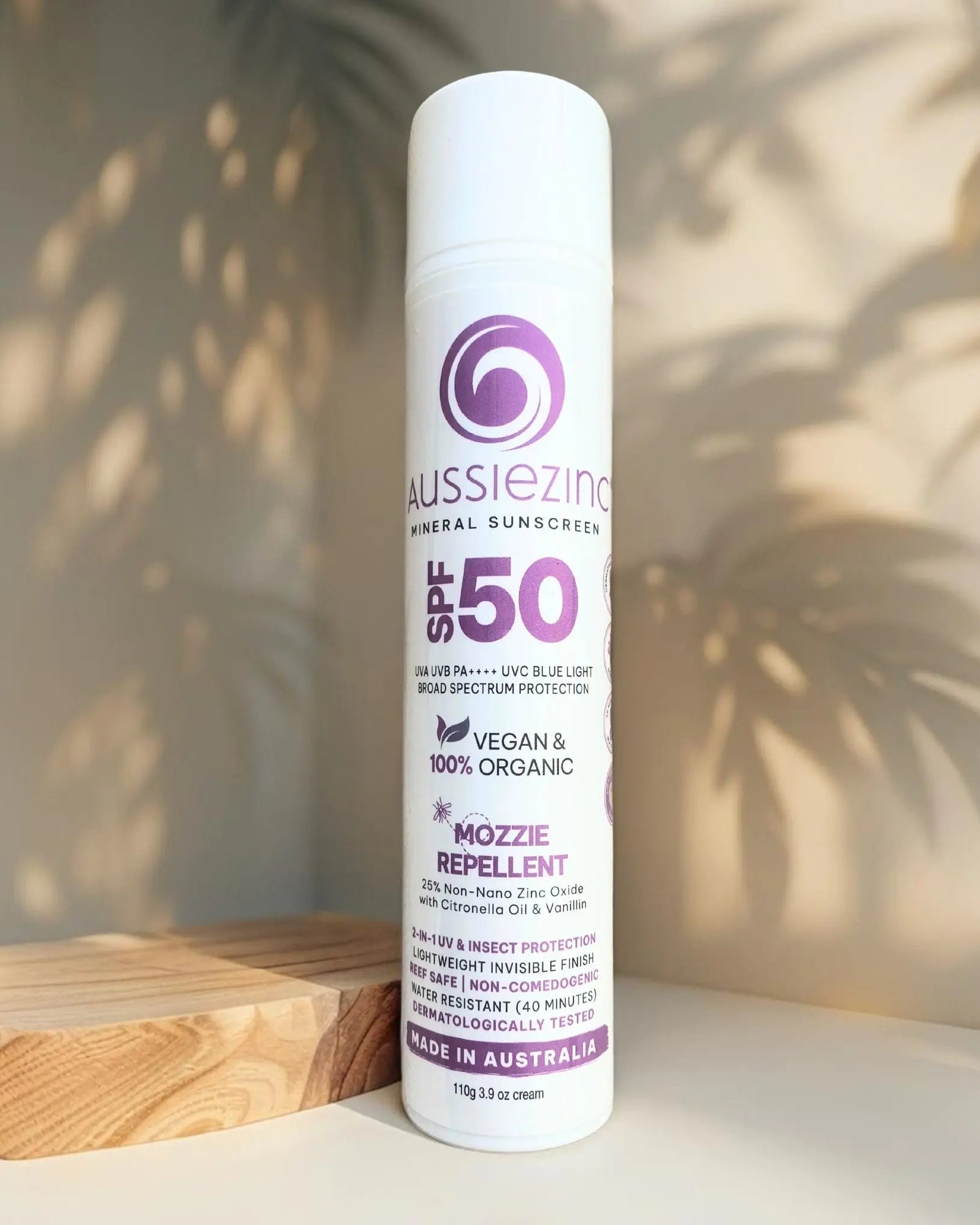 A bottle of AussieZinc 2-in-1 Mozzie Repellent SPF50+ Organic Mineral Zinc Sunscreen placed on a wooden surface with a shadow of a plant leaf on the bottle.